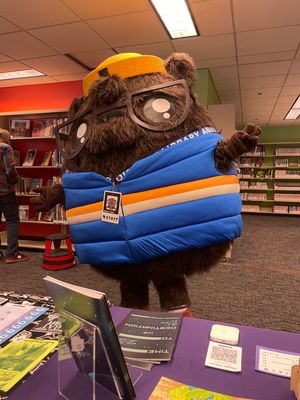 Akako G. Shins, the AADL mascot, standing in front of Maggie's table in the library and making a peace sign. Akako is a very round and fluffy groundhog mascot with glasses, a hat, a puffy vest and an AADL staff lanyard.