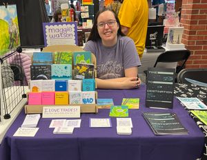 Maggie sitting at her table at A2CAF, smiling. On the table is a tiered stand with lots of her zines and a separate stack of copies of her Time to Destination comic.