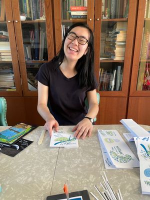 Maya sitting at a table surrounded by partly folded zines, smiling. 