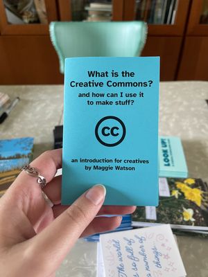 Maggie's hand holding up a zine titled 'What is the Creative Commons and how can I use it to make stuff? an introduction for creatives'